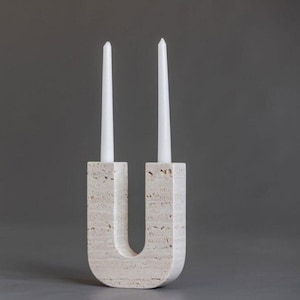 Puede incluir: Un candelabro de piedra beige en forma de U, con dos velas cónicas blancas. El candelabro tiene una superficie texturizada y un diseño moderno. Las velas son delgadas, añadiendo un toque de elegancia.