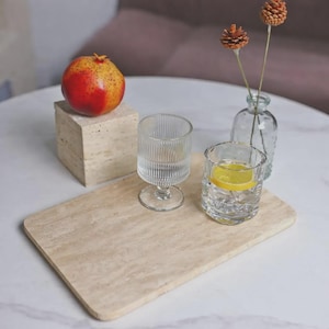 May include: A still life composition with a red and yellow pomegranate on a stone block, two clear drinking glasses, one with a lemon slice, and a small glass vase with dried flowers. The items are on a light-coloured rectangular tray.