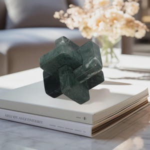 May include: A dark green marble sculpture in a geometric knot design. The sculpture is displayed on top of two white books. The books have text on the spine and cover. A vase of dried flowers is in the background.