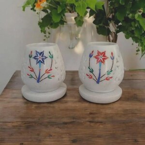 Peut inclure: Deux bougeoirs en pierre blanche avec des corps cylindriques et des bases circulaires. Chaque bougeoir présente un motif découpé et un dessin floral peint à la main. Celui de gauche affiche une fleur bleue, celui de droite une fleur rouge.