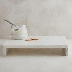 May include: A rectangular white marble serving board with a smooth surface. A clear glass bowl containing a creamy white dip sits on the board, accompanied by a silver spreader knife and two round, light brown crackers placed on the marble surface.