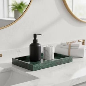 Puede incluir: Una bandeja rectangular de mármol verde oscuro que sostiene un dispensador de jabón negro mate con bomba y un recipiente cilíndrico de mármol blanco con estrías verticales lleno de bastoncillos, junto a toallas blancas plegadas.