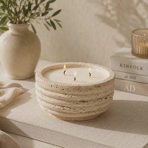 May include: A round, beige, textured stone candle vessel with three lit wicks and white wax. The exterior features horizontal ridges and a porous, natural finish. The candle is positioned on a flat, light-colored surface, creating a neutral aesthetic.