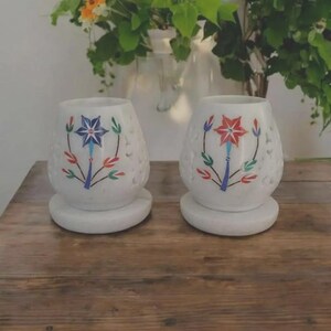 Op de afbeelding: Twee witte keramische kaarsenhouders met een ronde vorm en een getextureerd oppervlak. Elke houder heeft een handgeschilderd bloemmotief met een centrale stervormige bloem in blauw of rood, omringd door groene, rode en blauwe bladeren.