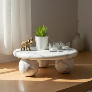 May include: A round white marble pedestal stand with three spherical marble legs. On top sits a small green succulent in a white ceramic pot, a gold-toned elephant figurine, and two clear glass tea light candle holders with white wax candles.