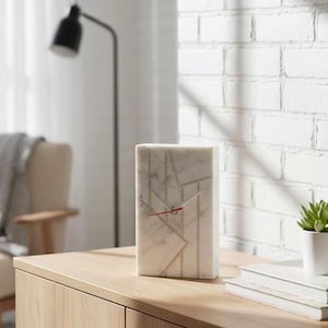May include: A rectangular white marble clock featuring a geometric gold-toned line pattern across its front surface. The clock has two silver-toned hour and minute hands and a thin red second hand centered on the marble face.