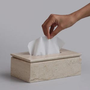 Puede incluir: Un dispensador de pañuelos rectangular de piedra beige. Una mano se extiende para sacar un pañuelo blanco. La caja tiene una parte superior plana con una abertura rectangular para el acceso a los pañuelos.