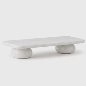 Puede incluir: Una tabla de servir de mármol blanco con una parte superior rectangular y dos soportes esféricos redondeados. La tabla tiene una superficie lisa y pulida y un patrón veteado natural. Ideal para servir aperitivos o exhibir artículos.