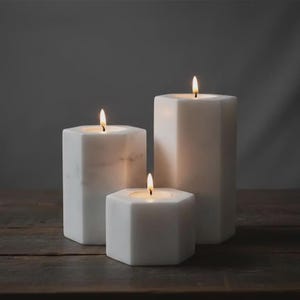 May include: Three white marble candles with a hexagonal shape and smooth finish. Each candle features a single lit wick with a warm flame. The candles are arranged in a group, with varying heights, showcasing the natural veining of the stone material.