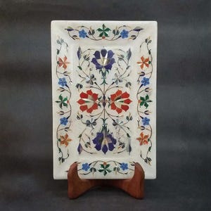 Puede incluir: Una bandeja rectangular de mármol blanco con intrincadas incrustaciones florales. El diseño incluye patrones simétricos de pétalos y enredaderas de piedra en colores rojo, azul, naranja y verde. La bandeja se apoya en un soporte de madera marrón.