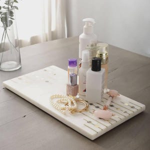 Könnte beinhalten: Ein rechteckiges Tablett aus weißem Marmor mit goldenen linearen Akzenten und kreisförmigen Einlagen. Auf dem Tablett stehen eine weiße Pumpspenderflasche, eine klare Flasche mit violettem Deckel, eine weiße Sprühflasche, eine quadratische Flasche, ein Perlenarmband und ein rosa Gesichtsroller.