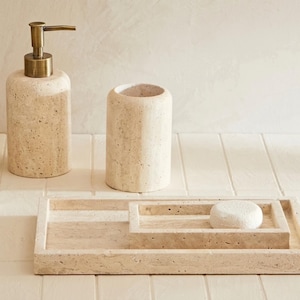 May include: A beige travertine bathroom set consisting of a cylindrical soap dispenser with a bronze-toned pump, a matching cylindrical toothbrush holder, and a rectangular tray holding a small, round, white textured soap bar.