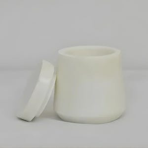 May include: A white cylindrical container with a slightly tapered shape and a matching circular lid placed beside it. The object features a smooth, solid-coloured surface with a matte finish, showing a clean, simple design with no additional patterns.