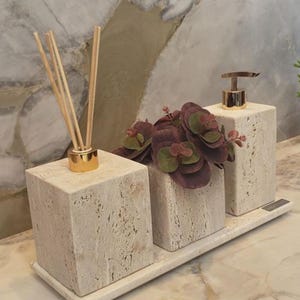 Puede incluir: Un juego de baño que presenta una bandeja con tres recipientes cúbicos con textura de piedra. Uno contiene un difusor con tapa dorada y varillas, otro tiene follaje falso morado y verde, y el tercero tiene un dispensador de jabón con tapa dorada.