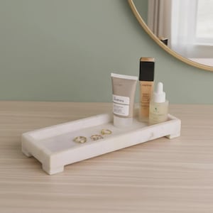 Pode incluir: Uma bandeja retangular de mármore branco com pés curtos, contendo três anéis dourados, um tubo de produto de cuidados com a pele The Ordinary, um frasco de base Lancôme preto e dourado e um pequeno frasco conta-gotas branco.