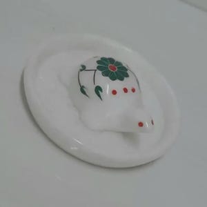 Może przedstawiać: Biała ceramiczna podstawka z wyrzeźbioną figurką żółwia pośrodku. Skorupa żółwia ozdobiona jest zielonym wzorem kwiatowym i małymi czerwonymi kropkami. Podstawka ma gładkie, błyszczące wykończenie i okrągły, płytki kształt.