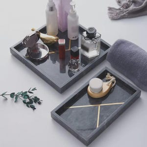 May include: Two dark gray marble-patterned trays with gold-colored geometric lines. The square tray holds various cosmetic bottles, a compact mirror, and a small crystal. The rectangular tray holds a wooden facial brush with a rope handle.