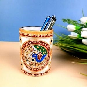 May include: A cylindrical white stone pen holder featuring a central circular inlay with a colorful peacock motif in blue, green, and orange. The rim and base are decorated with gold-toned bands and intricate red and gold patterns. Two pens are inside.