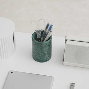 May include: A green marble pen holder with scissors, pens, and pencils. The cylindrical holder is filled with office supplies. A white speaker and a tablet are also visible on the white surface.
