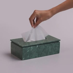 Puede incluir: Caja rectangular de pañuelos de mármol verde. Una mano saca un pañuelo blanco. La caja tiene tapa y una abertura rectangular para dispensar pañuelos. El mármol presenta un veteado natural.