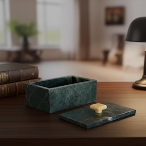 May include: A rectangular, dark green marble box with a matching lid. The lid is off, revealing the box's interior. The lid has a square, cream-colored knob. The box and lid sit on a dark wooden surface, with a lamp and books in the background.