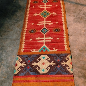 Puede incluir: Una alfombra pasillera larga y rectangular con un campo central de color rojo vibrante que presenta motivos geométricos repetitivos de diamantes y cruces en turquesa, blanco y marrón. Los extremos tienen bordes marrones con flecos.