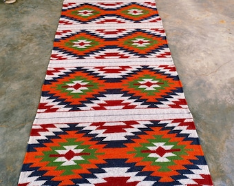 May include: A long, rectangular rug with a geometric pattern in orange, red, green, blue, and white. The rug has a fringed edge and is likely made of woven fabric. The design features diamond and zigzag shapes.