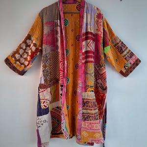 May include: A long-sleeved robe featuring a patchwork design made from various fabrics in shades of orange, pink, yellow, and white. The garment includes a bright pink trim along the front opening, two front pockets, and a matching fabric tie belt.