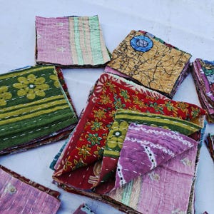 May include: A collection of folded fabric squares featuring various patterns and colors, including floral, geometric, and abstract designs in shades of pink, green, red, yellow, and blue, all finished with visible white running stitch quilting.