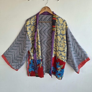 May include: A patchwork kimono jacket featuring a mix of patterns and colors. The garment displays sections of black and white chevron, yellow floral prints, and vibrant red panels with blue flowers. It has wide sleeves and a purple trim border.