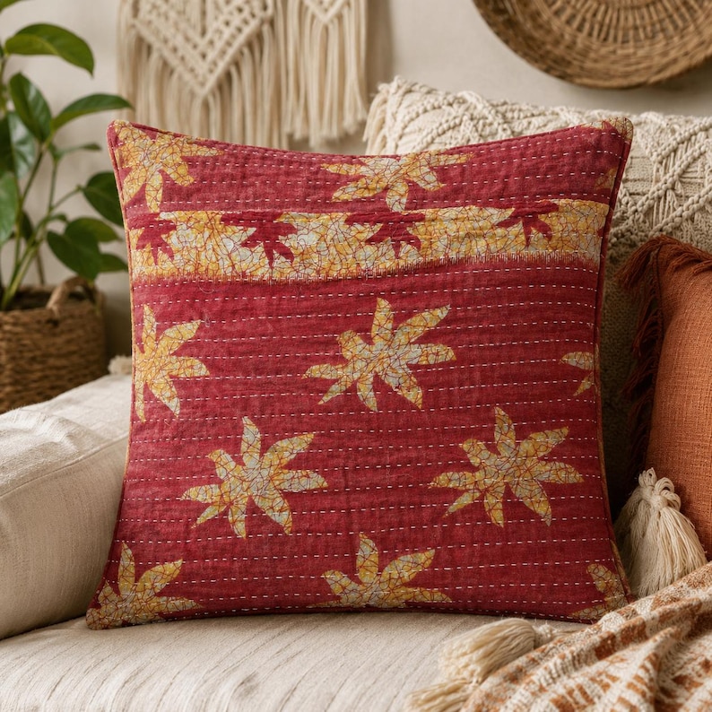 May include: A square throw pillow featuring a deep red base color with a repeating pattern of yellow and white star-shaped leaf motifs. The fabric displays visible white horizontal running stitch quilting details and a horizontal band.