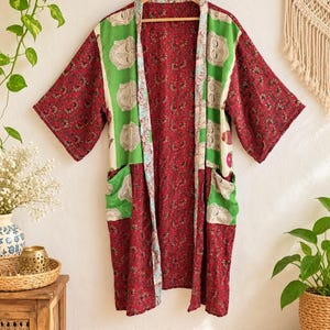 May include: A long-sleeved kimono-style robe featuring a deep red fabric with a dense floral pattern. The front panels and pockets are accented with a contrasting light green fabric decorated with a repeating line-art pattern of faces.