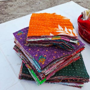 May include: A stack of rectangular fabric swatches featuring various colors and patterns, including bright orange with white leaf motifs, deep purple with geometric designs, and dark green with white stitching throughout the layered pieces.