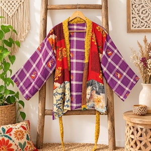 Könnte beinhalten: Eine Patchwork-Kimonojacke aus verschiedenen Stoffbahnen mit violettem Karomuster, roten Blumenmustern und blauen botanischen Motiven. Das Kleidungsstück hat weite Ärmel, einen gelben Kragen und zwei lange gelbe Bindebänder.