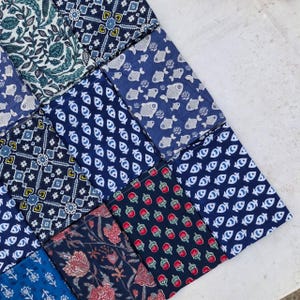 Puede incluir: Una colección de cuadrados de tela doblados con varios patrones, incluyendo peces azules y blancos, motivos florales, formas geométricas y pequeñas bayas rojas sobre un fondo azul oscuro, dispuestos en cuadrícula sobre una superficie blanca.