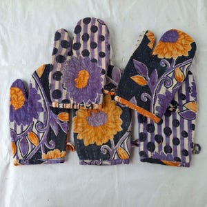 May include: A set of three oven mitts featuring a quilted texture with visible stitching. The fabric displays a pattern of large orange flowers, purple floral motifs, and black polka dots on a cream background, arranged in various block designs.