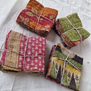 May include: Four stacks of folded fabric squares featuring various patterns and colors, including red, green, yellow, and pink. Each stack is tied with thin white string and displays visible white running stitch embroidery throughout the material.