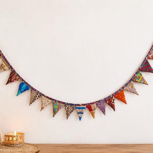 Könnte beinhalten: Eine dekorative Wimpelkette bestehend aus einer Reihe dreieckiger Stofffahnen in verschiedenen Farben und Mustern, darunter florale, geometrische und gestreifte Designs, aufgehängt an einer dunklen Stoffkordel mit Schlaufen an beiden Enden.