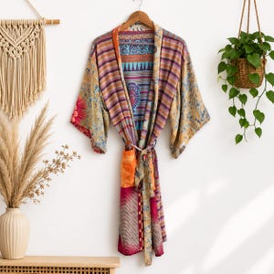 May include: A long-sleeved kimono robe featuring a patchwork of vibrant patterns including stripes, floral motifs, and geometric designs in shades of orange, magenta, blue, and gold. The garment includes a matching fabric tie belt at the waist.
