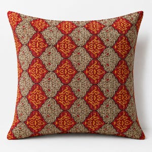 May include: A square throw pillow featuring a repeating pattern of red and beige quatrefoil shapes. The red sections contain yellow swirling paisley motifs, while the beige sections feature intricate brown floral patterns with visible white stitching.