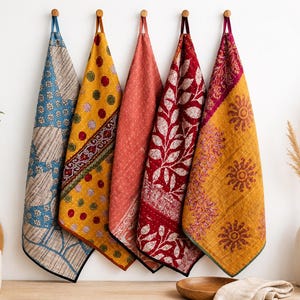 Könnte beinhalten: Fünf rechteckige Stoffhandtücher hängen in einer Reihe, jedes mit unterschiedlichen Mustern und Farben, darunter Blau mit Blumenmotiven, Gelb mit roten Punkten, einfarbiges Koralle, Rot mit weißen Blattdrucken und Gelb mit sonnenartigen Kreismustern.