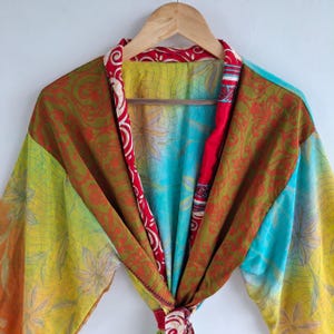 Könnte beinhalten: Ein leichter Kimono-Bademantel mit einem lebendigen Patchwork-Design und Blumenmustern in Gelb-, Türkis- und Orangetönen. Das Kleidungsstück verfügt über einen kontrastierenden rot-weißen Saum entlang des Kragens und der vorderen Öffnung.