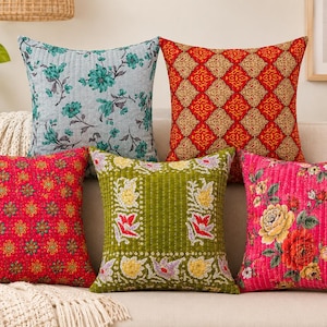 May include: Five square throw pillows with varied patterns. Top left is light blue with teal flowers. Top right is red and gold geometric. Bottom left is red with small floral motifs. Bottom center is olive green with floral borders. Bottom right is pink with large roses.
