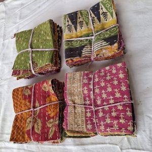 May include: Four stacks of folded fabric squares featuring various patterns and colors, including green, yellow, red, and pink. Each stack is secured with a thin white string and displays visible white running stitch embroidery throughout.