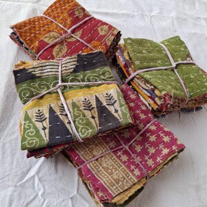 May include: Three folded stacks of multi-colored fabric squares featuring various patterns including floral, geometric, and tree motifs. Each stack is secured with thin, light-colored string and displays visible white running stitch embroidery.