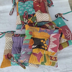 May include: A collection of square seat cushions featuring a patchwork design of various colorful fabrics with intricate kantha stitching. Each cushion includes fabric ties at the corners and displays a mix of floral, geometric, and abstract patterns.