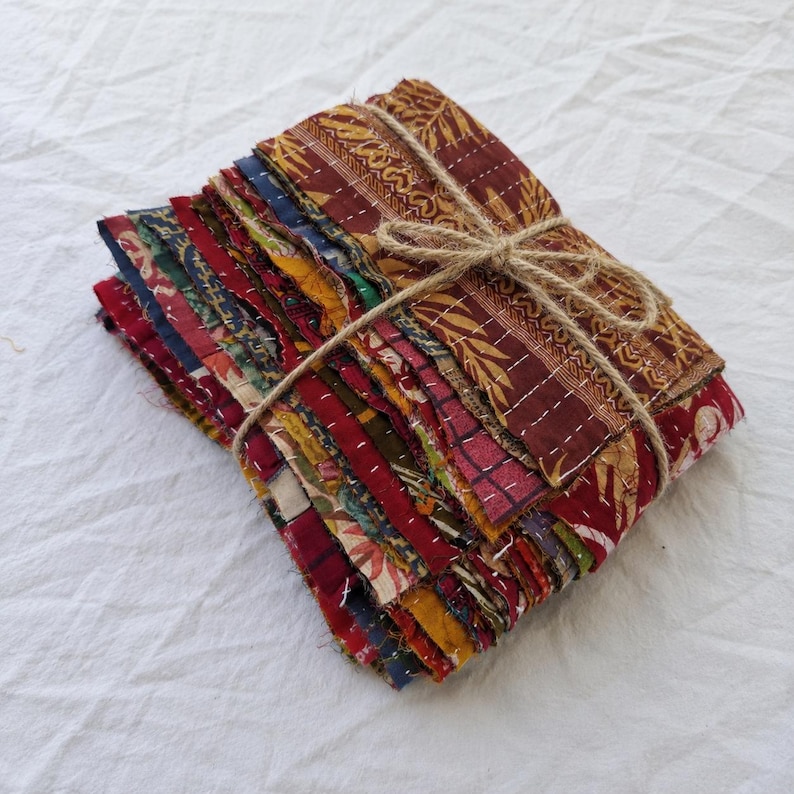 May include: A stack of rectangular fabric swatches featuring various patterns and colors including deep red, gold, green, and blue. The fabric pieces are layered and secured together with a simple brown twine tied in a bow across the center.