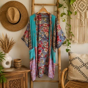 May include: A lightweight open-front kimono featuring a central panel with a floral pattern in shades of red, white, and blue on a grey background. The garment includes turquoise and lavender side panels, wide sleeves, and two front pockets.
