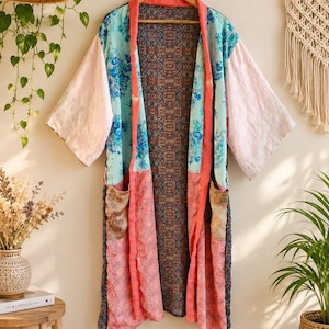 Könnte beinhalten: Ein langer, vorne offener Kimono mit Patchwork-Design, bestehend aus verschiedenen Blumen- und geometrischen Mustern in Hellblau, Rosa und Dunkelgrau. Das Kleidungsstück hat weite Ärmel, zwei Vordertaschen und einen korallenfarbenen Saum.