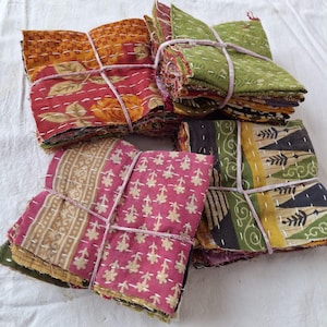 May include: Bundles of colorful fabric squares, each tied with pink string. The fabrics feature floral and geometric patterns in shades of red, green, pink, and yellow. White stitching accents the edges of the squares, creating a patchwork effect.